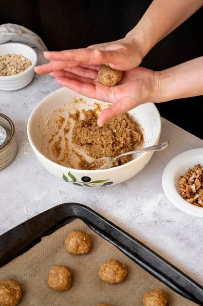 A close-up shot of a person's hands preparing a batch of homemade energy bites. The ingredients, including rolled oats, chia seeds, honey, and almond butter, are neatly arranged around a mixing bowl. The focus is on the hands rolling the mixture into small bite-sized balls.