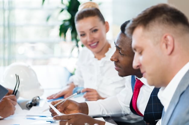 A diverse group of professionals sitting around a conference table, reviewing documents and discussing funding opportunities related to the infrastructure bill. Laptops and charts are displayed, showcasing project proposals and budget allocations.